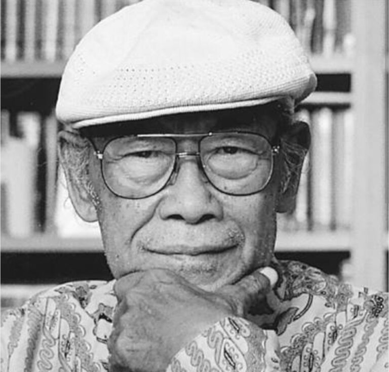 Older man wearing a white knit cap and glasses, resting his chin on his hand in a library setting (black-and-white photo).